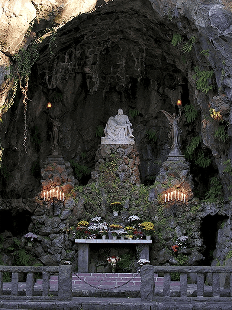 Peaceful shrine with statues, candles, and flowers within a rocky cave.