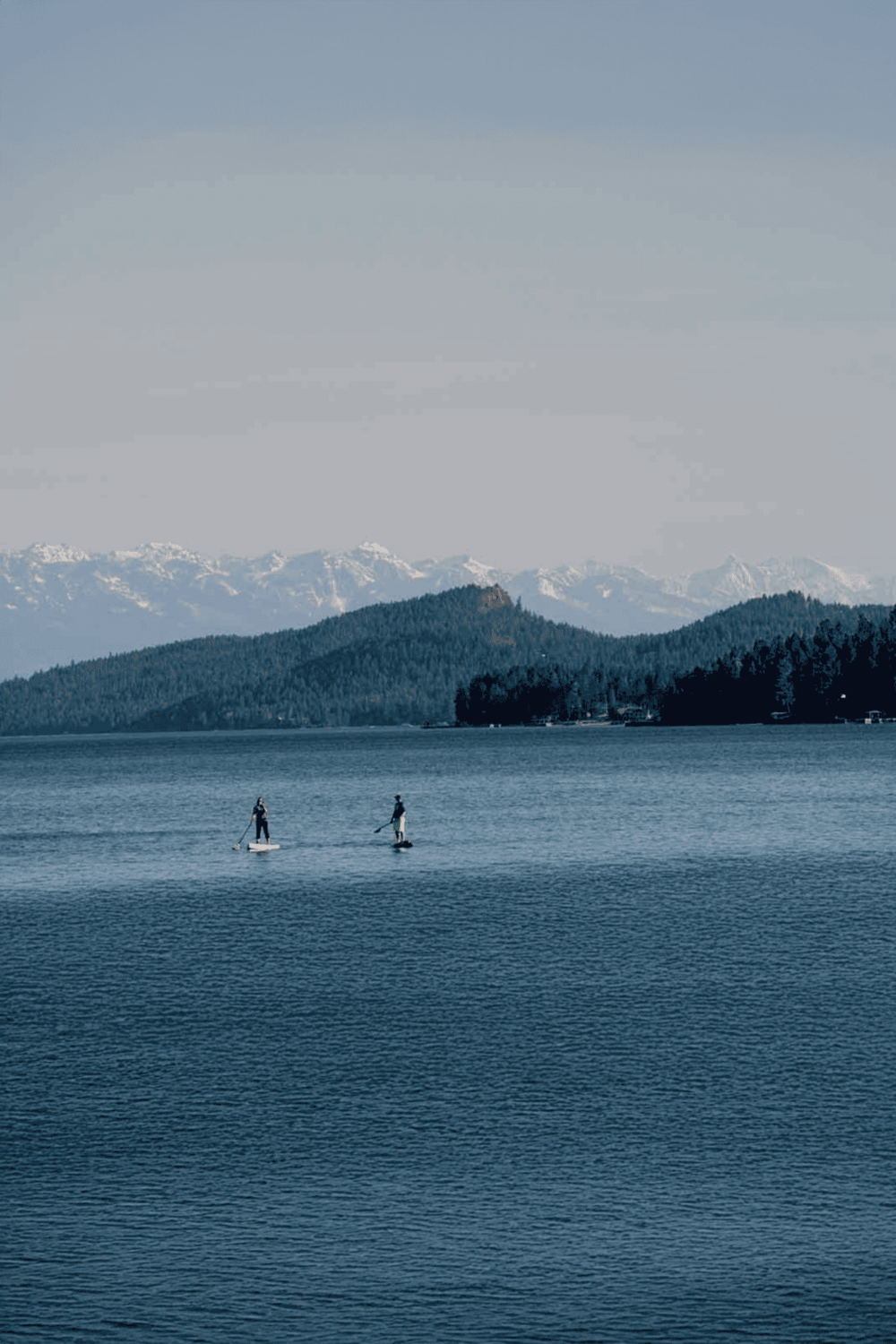 Serene lake with paddleboarders, lush forested hills, and snow-capped mountains in the background, perfect for outdoor adventure and nature exploration.