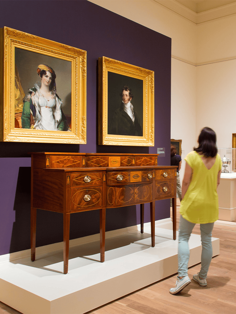 Antique wooden sideboard in art museum with portrait paintings overhead.