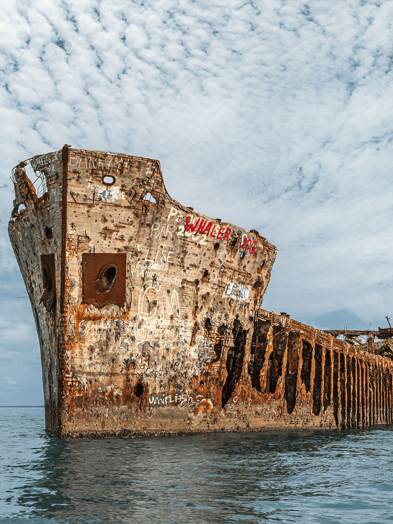 Rusty shipwreck on the ocean with graffiti on its sides and a cloudy sky in the background.