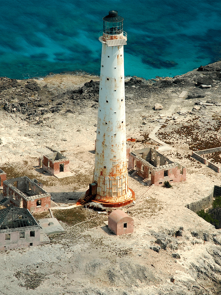 Lighthouse on rocky coast with abandoned buildings and ocean view in the background.
