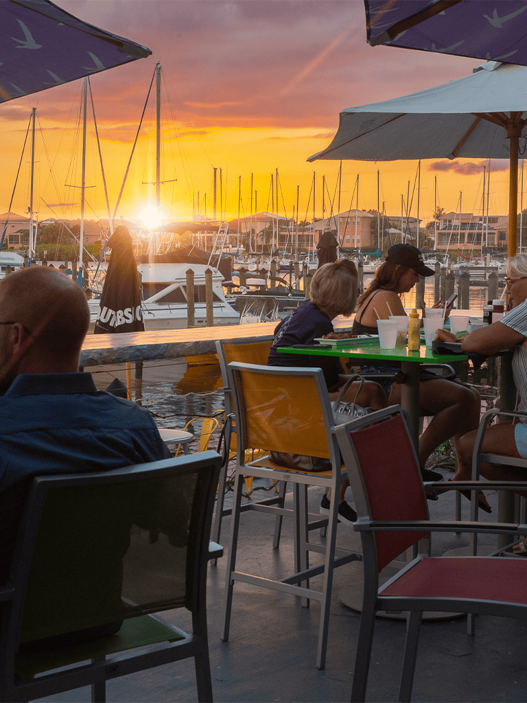 Sunset at the marina with people dining, boats, and vibrant sky, perfect for waterfront dining and relaxing views.