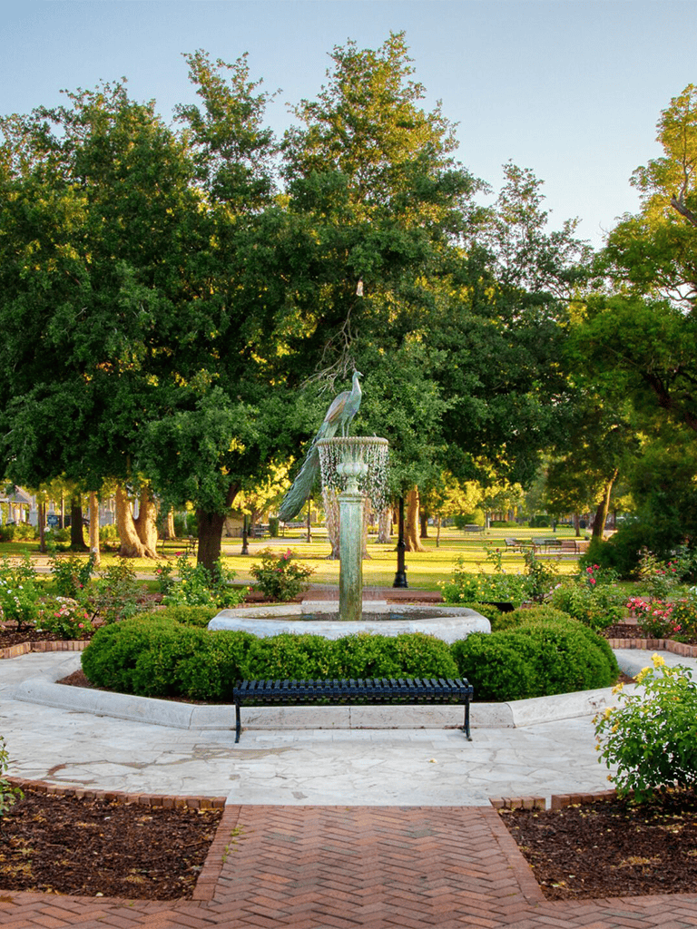 Colorful park fountain with a peacock statue surrounded by green trees and blooming flowers.