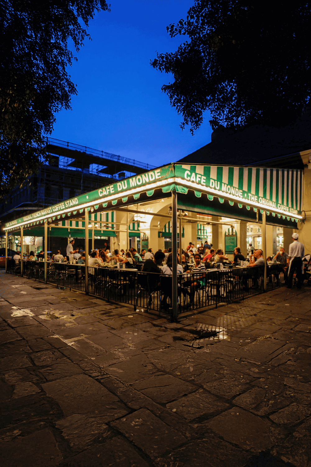 Cozy outdoor coffee shop at dusk with people dining under the green and white striped awning, vibrant city atmosphere, and night sky.