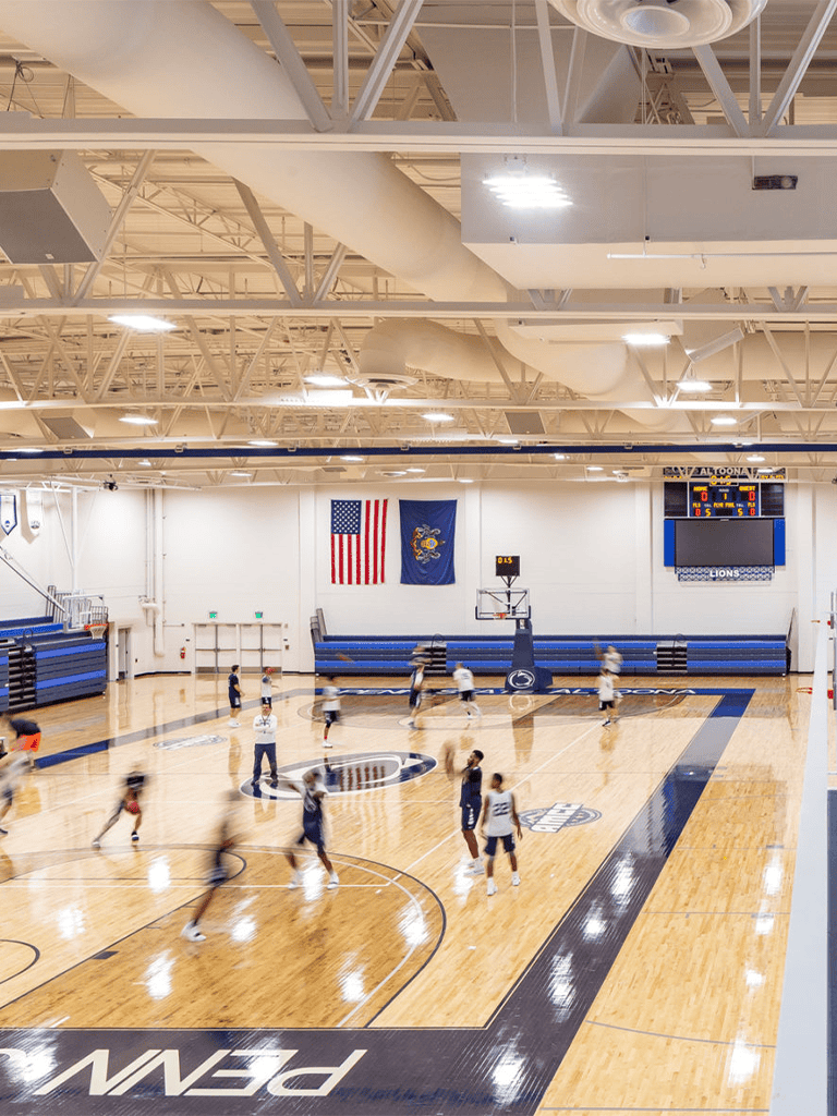 Indoor basketball gym with players practicing, American and state flags, and digital scoreboard.