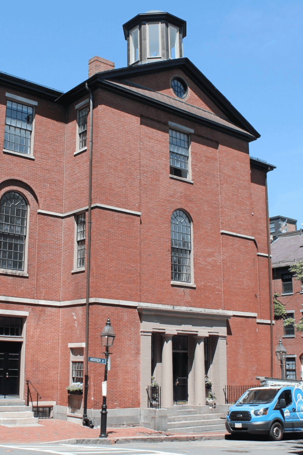 Historic red brick building with large windows and classical columns, located at a city intersection.