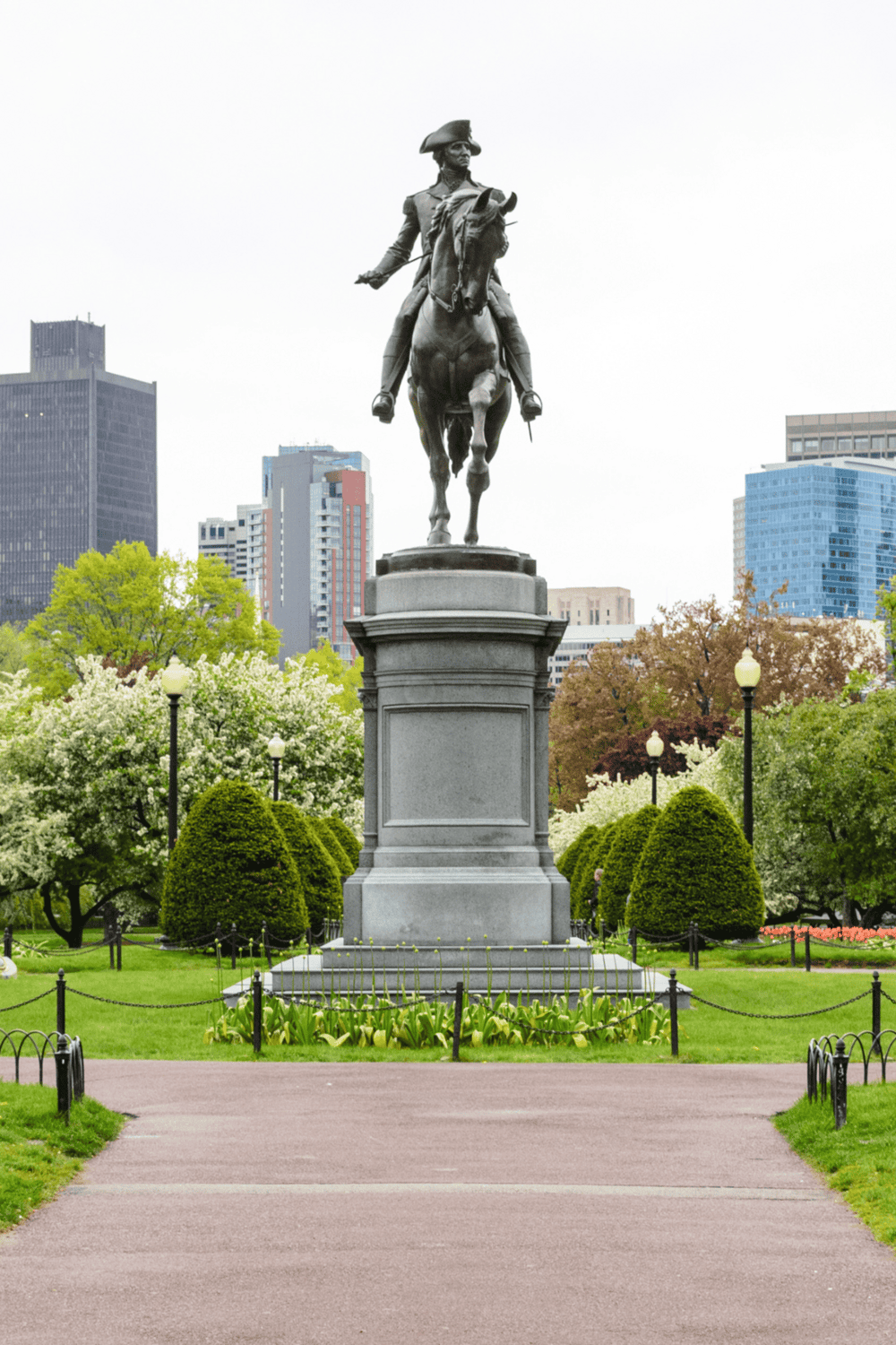 Bronze statue of a historical figure on horseback in a city park with lush greenery, modern buildings, and blooming trees.