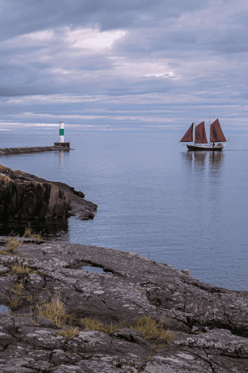 Serene coastal scene with a ship featuring red sails sailing near a lighthouse on a cloudy day.