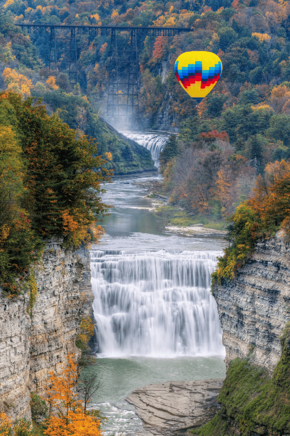 Vibrant autumn landscape with waterfall and hot air balloon over scenic gorge.