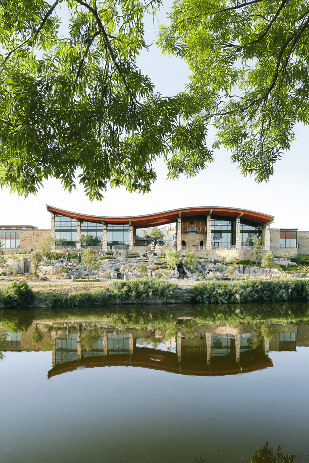 Contemporary building with large glass windows overlooking a pond, surrounded by greenery.