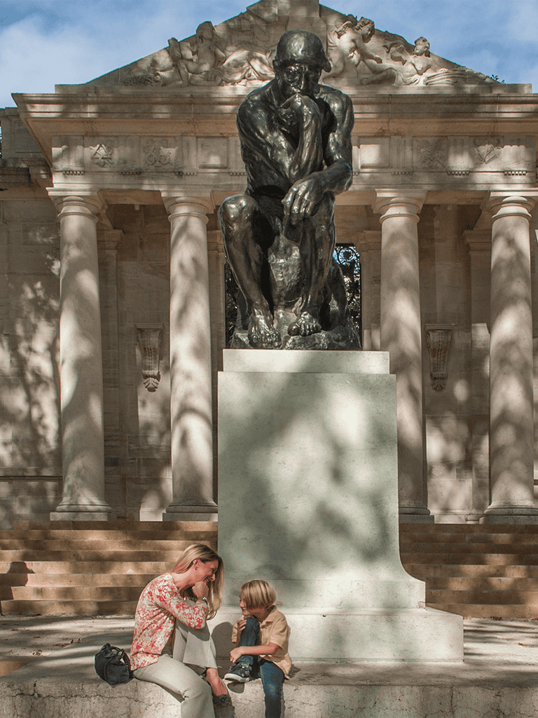 Pensative statue of a thinker at an outdoor monument with two children sitting nearby.