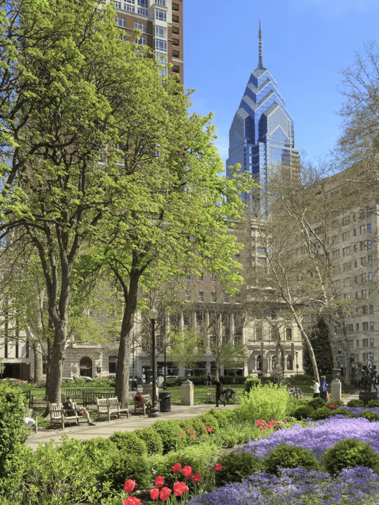 Lush city park with benches, colorful flowers, and towering skyscrapers in the background.