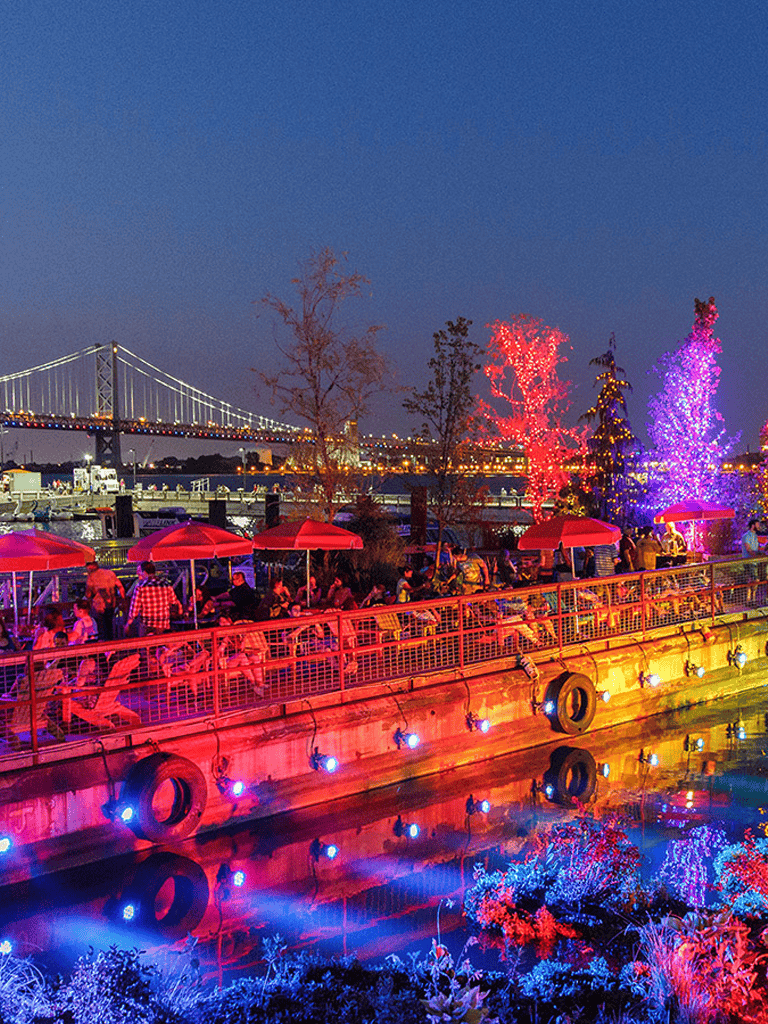 Colorful waterfront at night with illuminated trees, bridge, and people enjoying outdoor dining and nightlife.