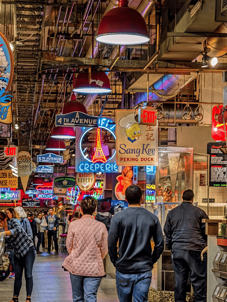 Vibrant indoor market with neon signs, diverse food vendors, and bustling shoppers, offering a lively dining and shopping experience.