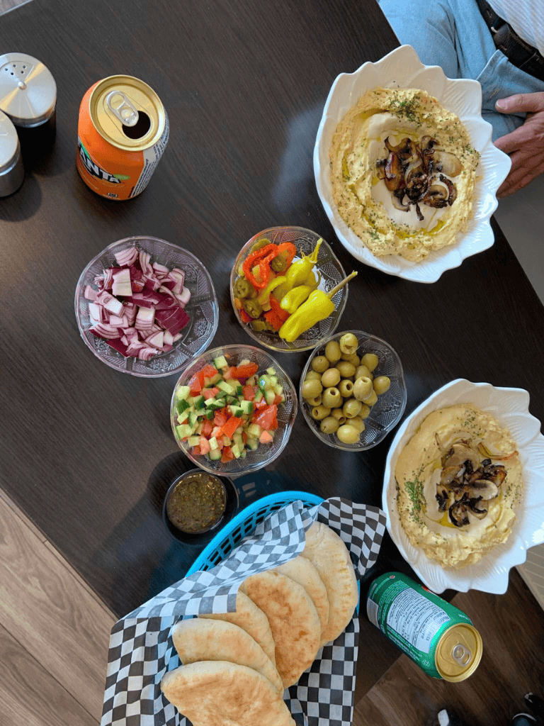 Fresh Mediterranean food with hummus, pita bread, salads, pickles, and canned drinks. Perfect for sharing and enjoying with friends.