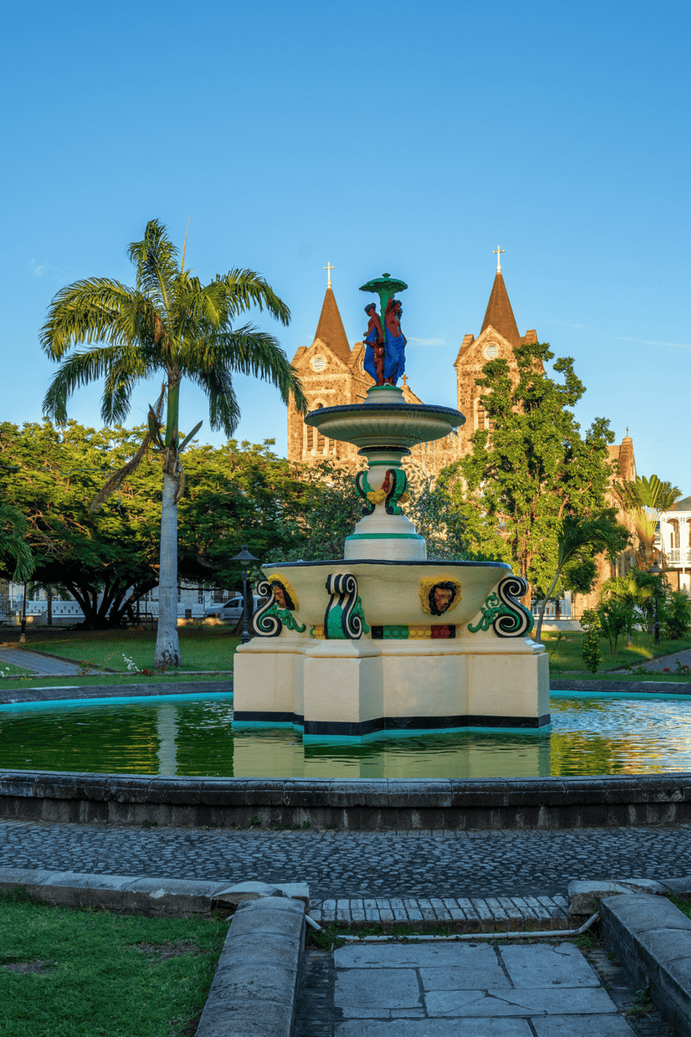 Colorful town square fountain with church in background, outdoor public space, tropical trees, scenic cityscape.