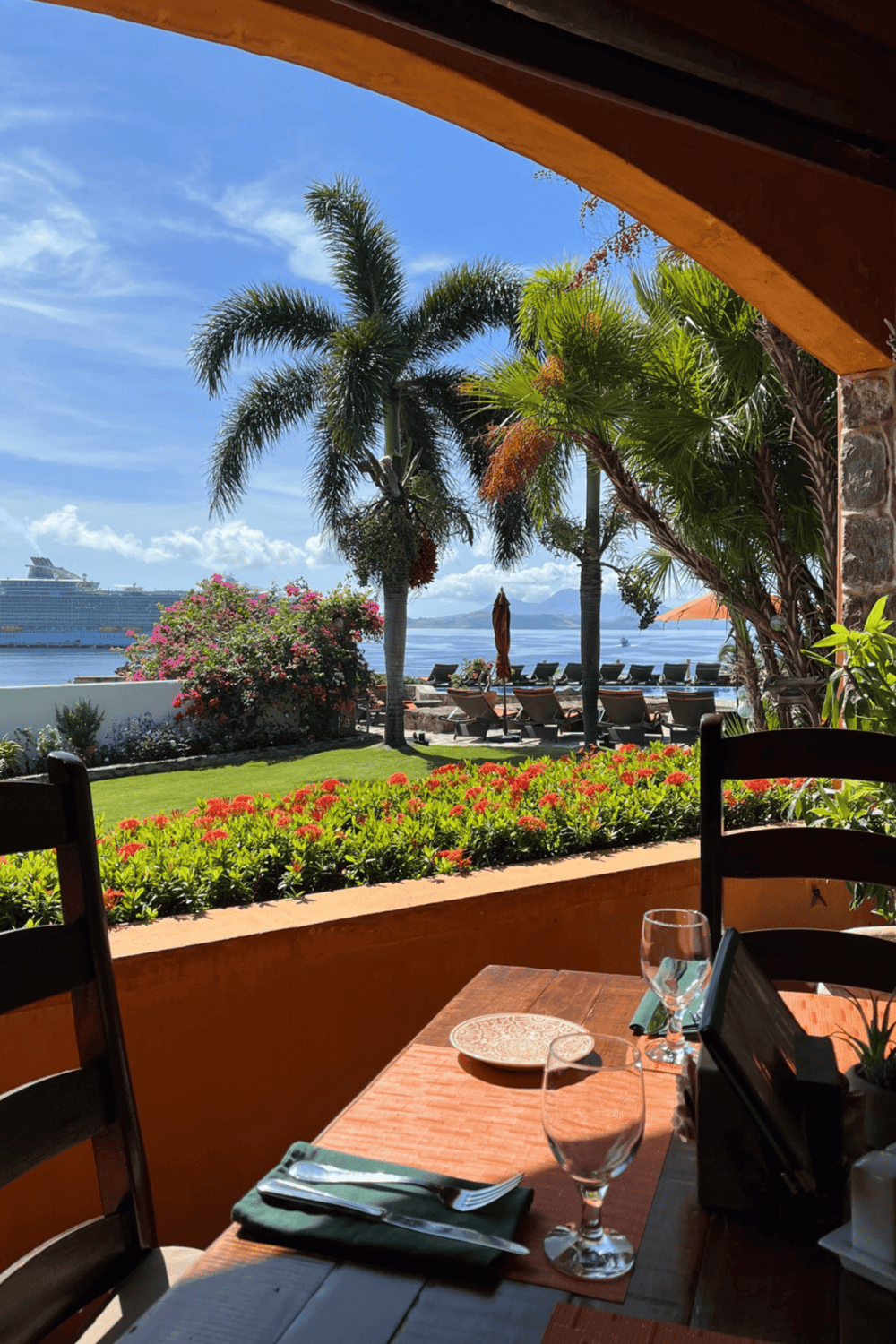 Vibrant tropical garden view with ocean, cruise ship, palm trees, and seaside lounge chairs.