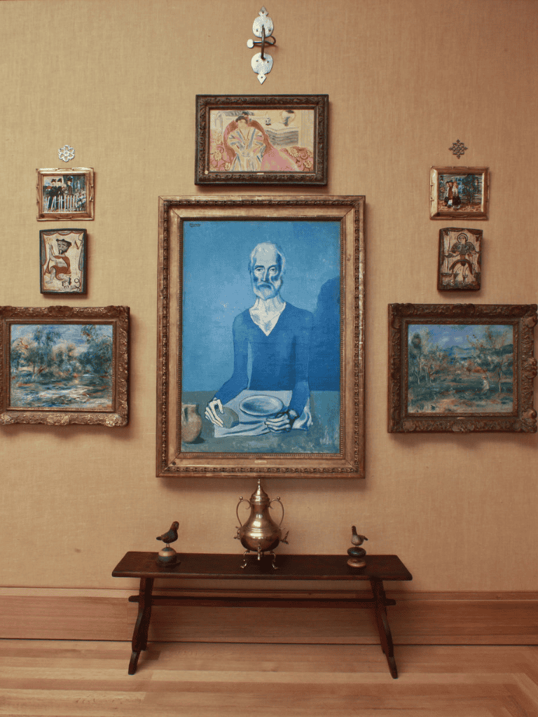 Old man portrait painting with multiple framed artworks on beige wall, vintage decor, and wooden bench.