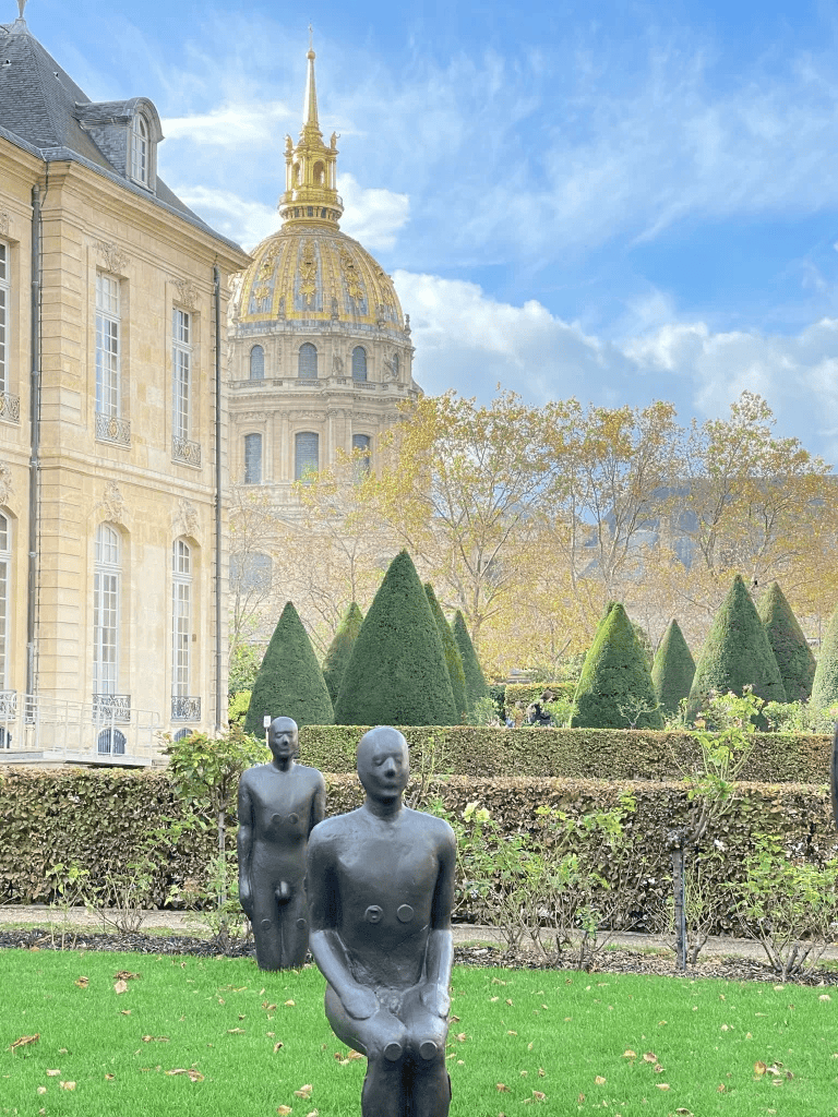Modern outdoor sculpture art featuring two abstract human figures in front of historic European architecture and manicured garden.