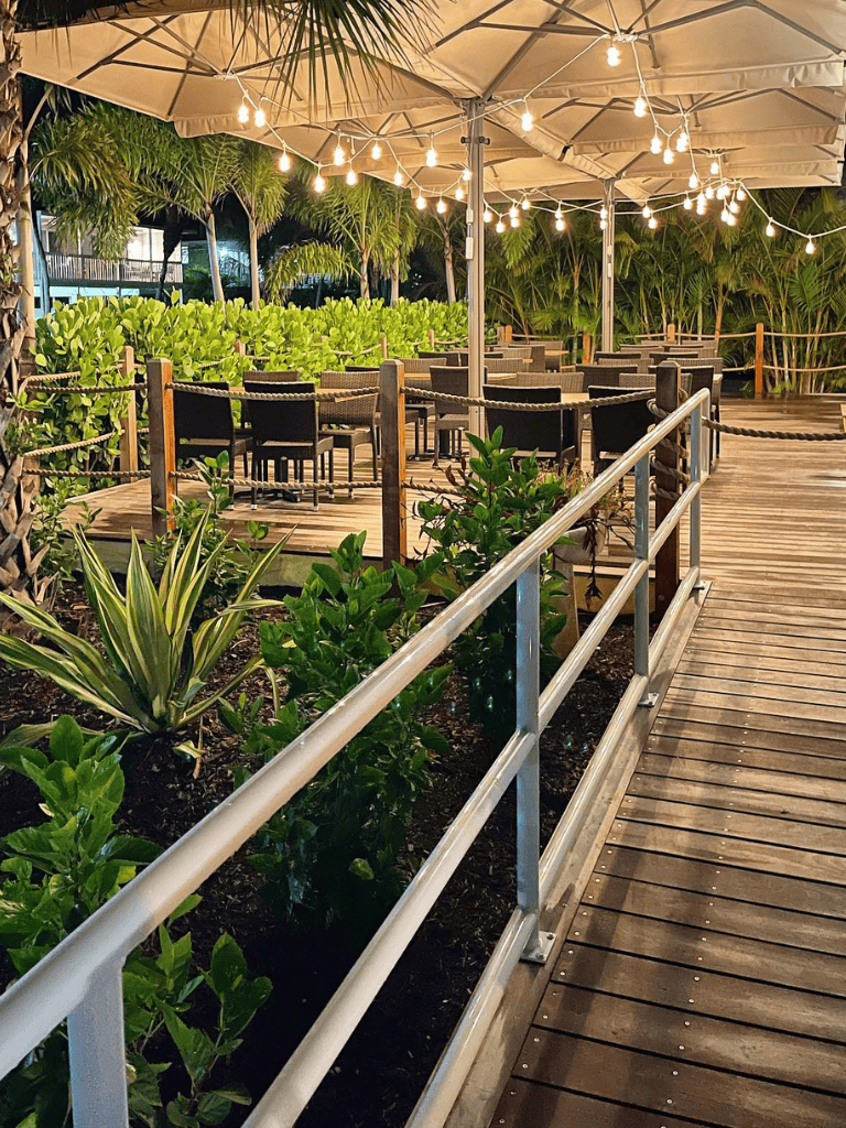 Outdoor dining area with string lights, umbrellas, lush greenery, and wooden flooring at QuestForDirections.