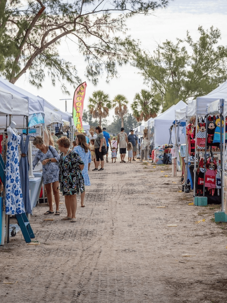 Colorful outdoor market with white vendor tents and shopping visitors, palm trees, and blue sky.