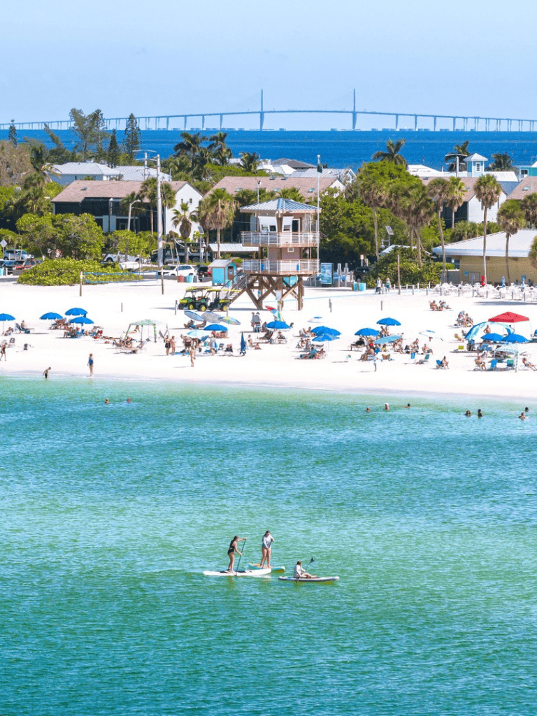Bright sunny beach with umbrellas, lifeguard tower, and paddleboarders on turquoise water.