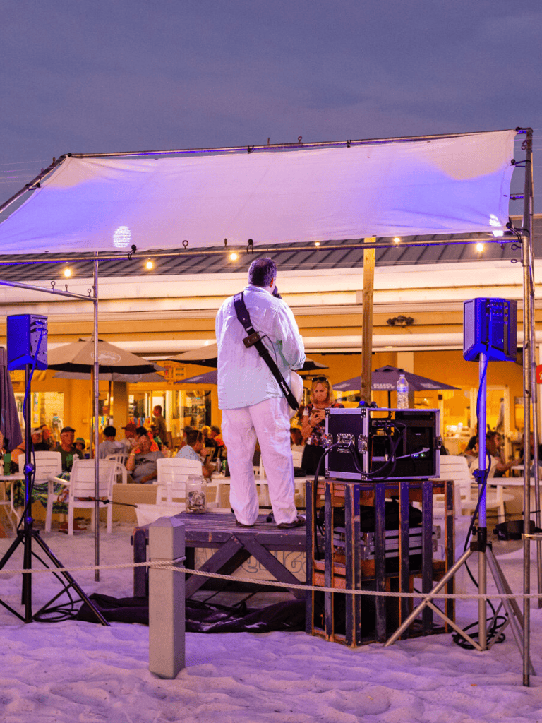 Live music performance on beach at sunset, people enjoying entertainment and drinks, ocean in background, lively beach party scene.