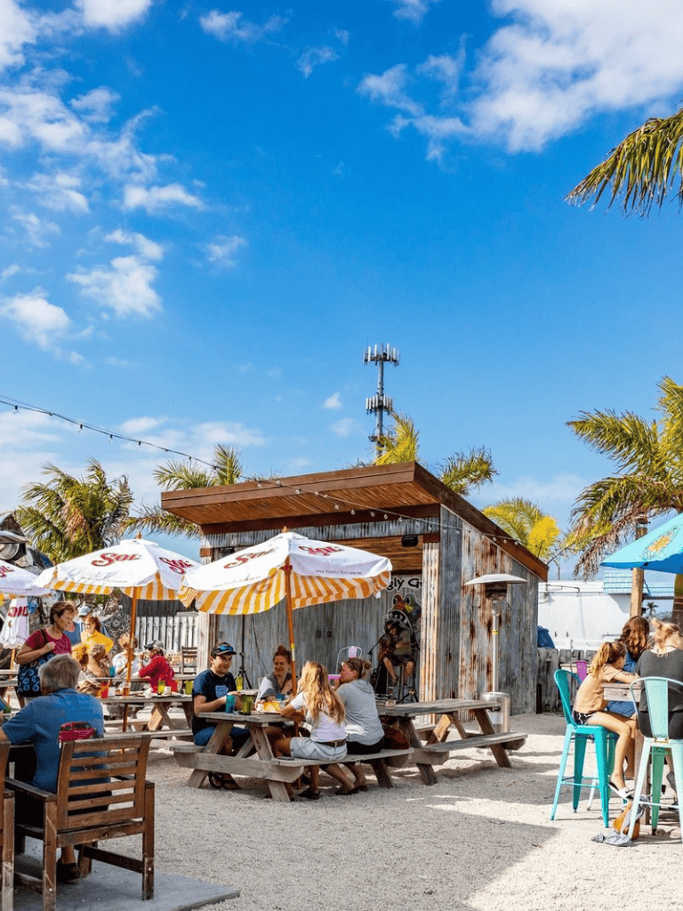 Outdoor dining area with picnic tables, umbrellas, and people enjoying food under sunny skies.