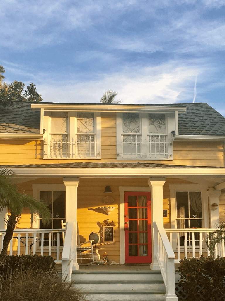 Charming yellow house with red door, white shutters, and porch, coastal style with tropical surroundings.