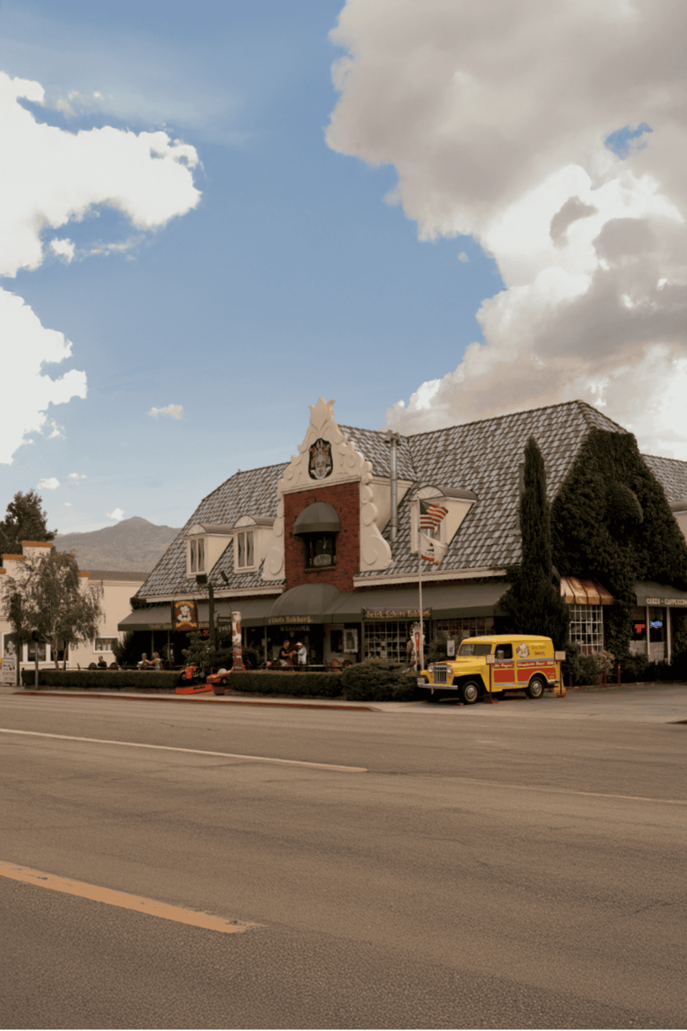 Charming European-style bakery and cafe storefront with vintage vehicle, outdoor seating, and scenic mountain backdrop.