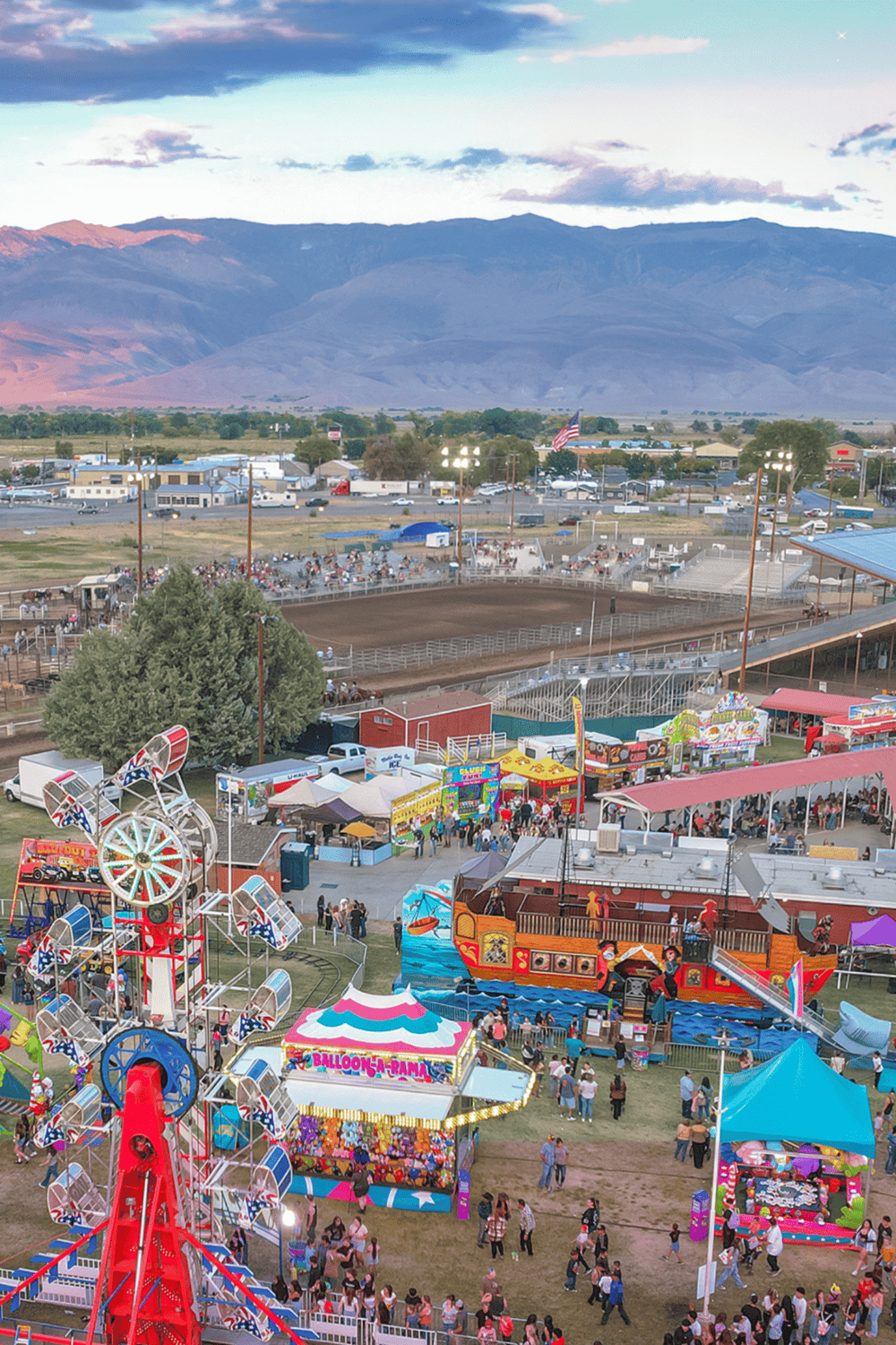 Colorful county fair with rides, games, and crowds, set against mountain background.