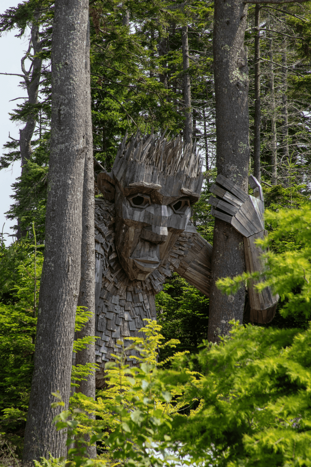 Wooden forest sculpture of a man in wilderness, nature art installation, outdoor wooden sculpture in a forest setting, tall trees surrounding the art piece.