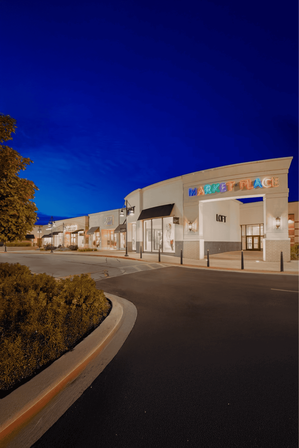Modern shopping center at dusk with lit storefronts and colorful "Market Place" sign.