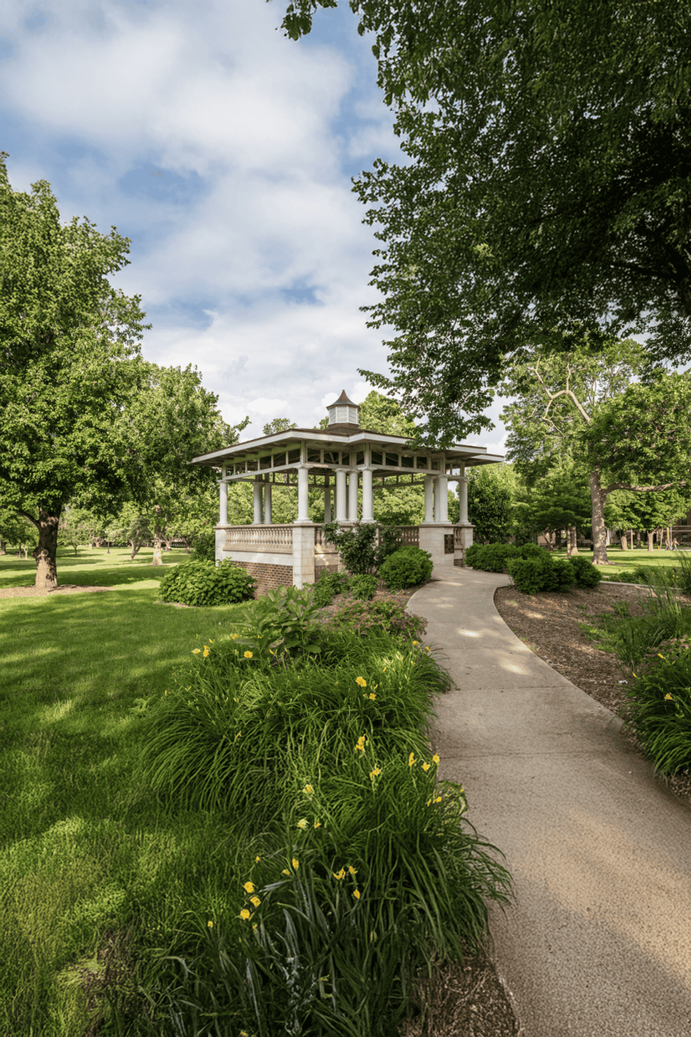 A picturesque park gazebo surrounded by lush greenery and vibrant flowers under a partly cloudy sky.