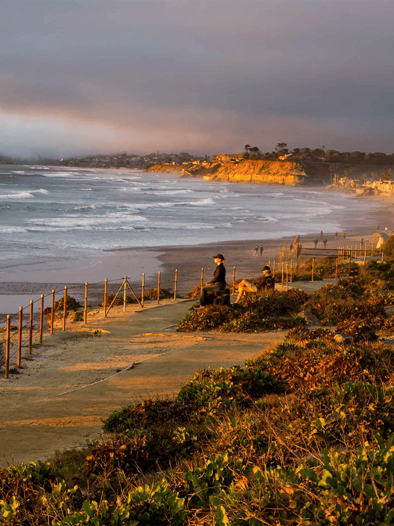Sunset coastal view with people on a seaside walkway, overlooking ocean waves and cliffs, inspiring sunset beach travel destinations.