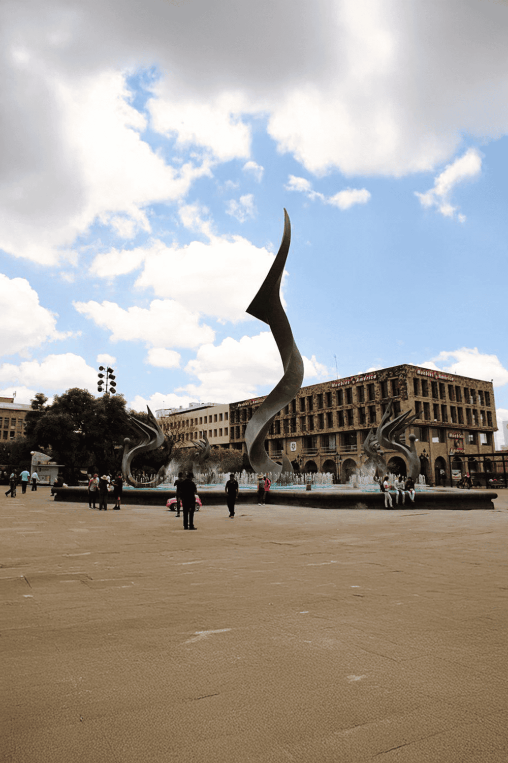 Contemporary sculpture fountain in the city square with modern buildings and people, popular tourist attraction.