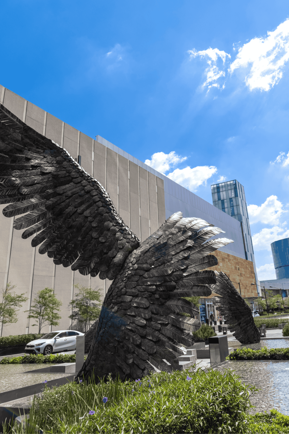 Bird sculpture art installation in modern cityscape with skyscrapers, greenery, and clear blue sky.