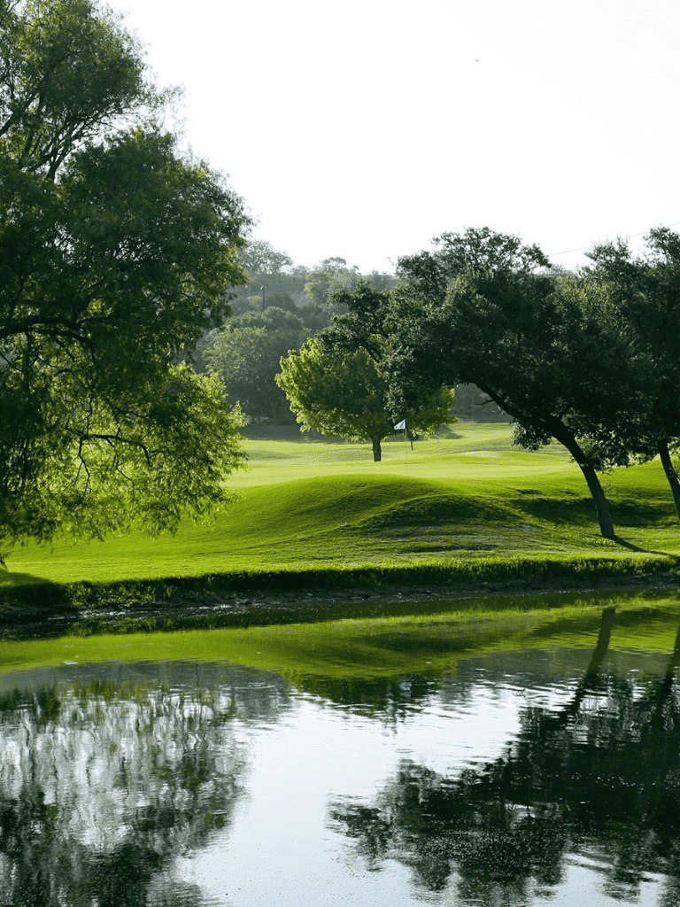Lush green golf course with water hazard and mature trees in bright sunlight.