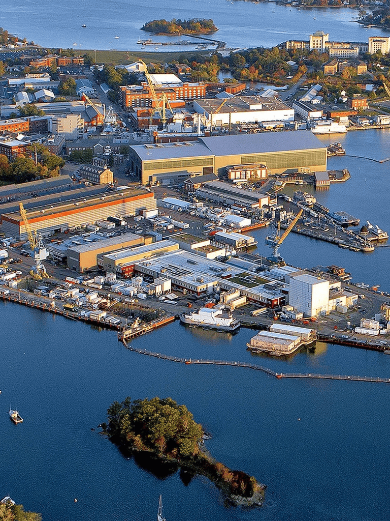 Modern industrial harbor with warehouses, cranes, boats, and docks in a coastal cityscape.