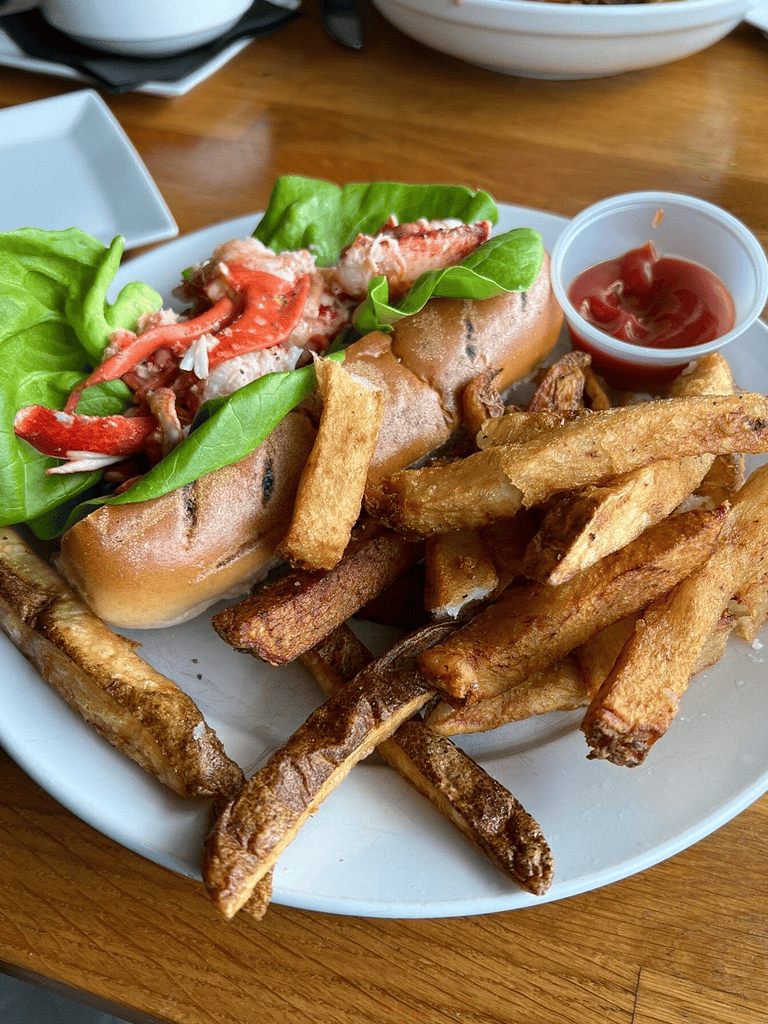 Juicy lobster roll with fresh greens, crispy fries, and ketchup on a white plate.