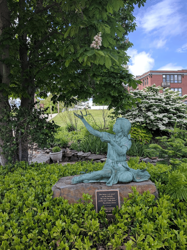 Child statue reaching for tree in a garden, lush greenery, peaceful outdoor scene with building in background.