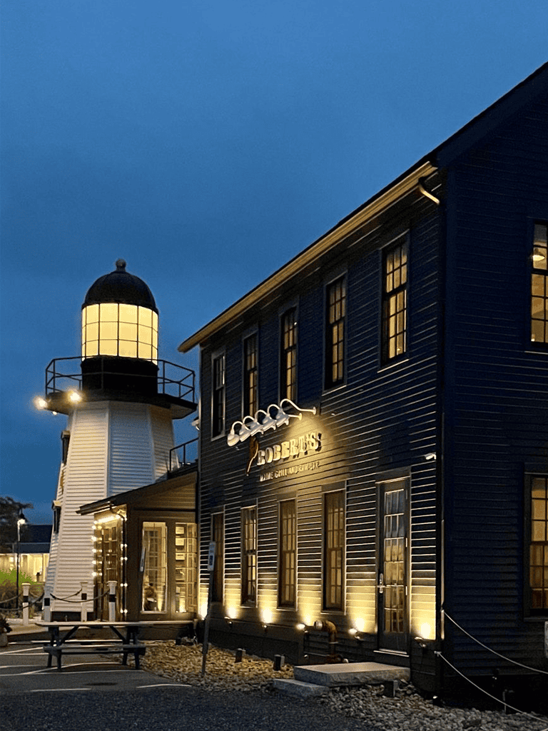 Lighthouse and coastal restaurant at dusk, illuminated exterior, inviting seaside dining experience.