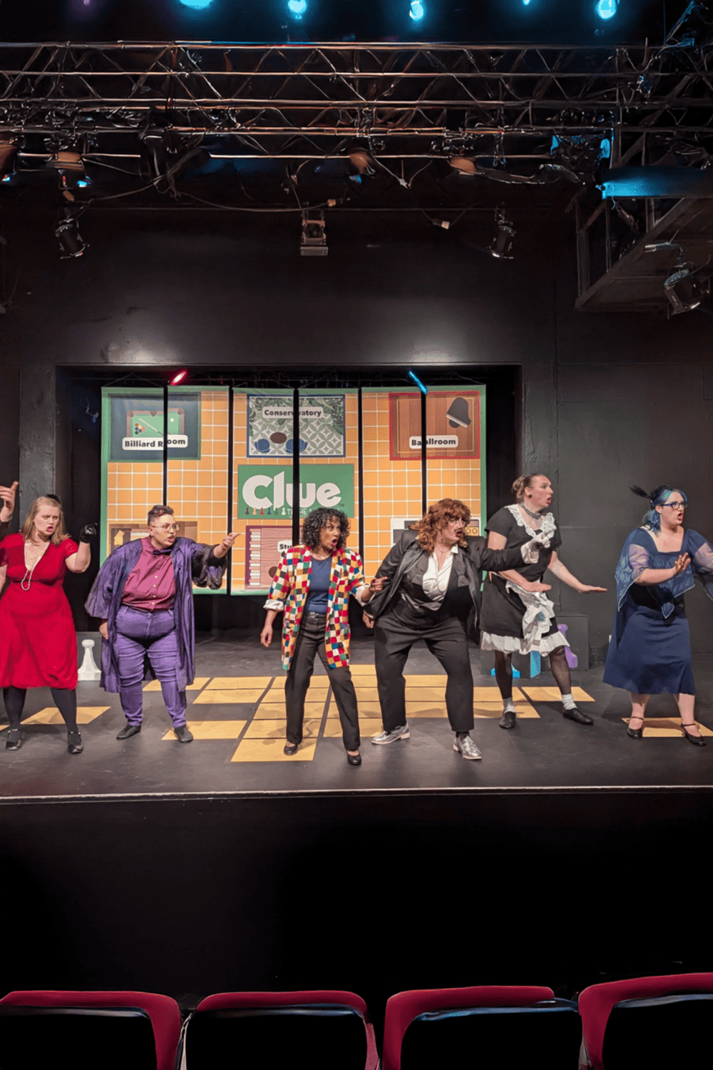 Diverse group of actors performing a stage play with vibrant set design and costumes on a theatrical stage.