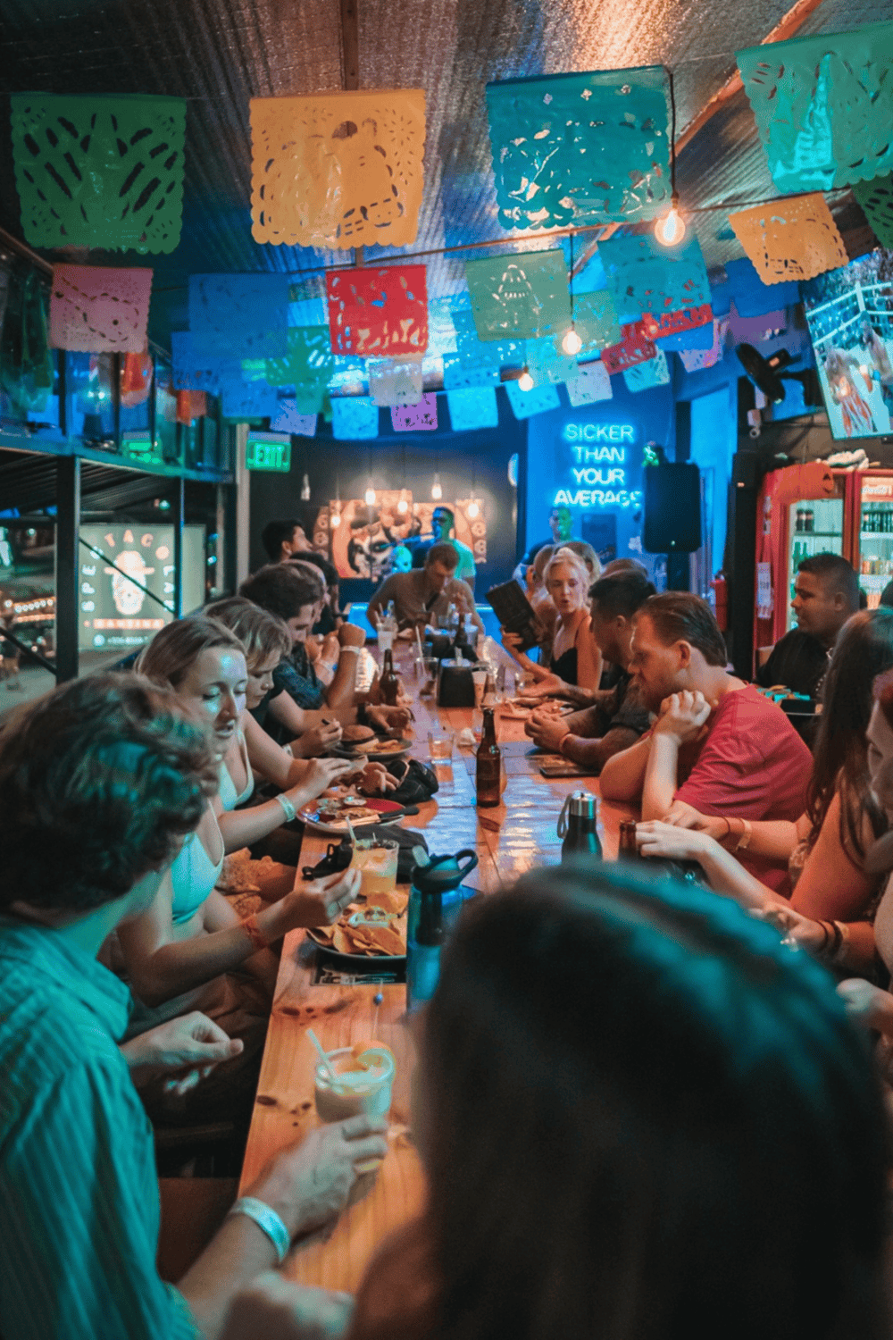 A vibrant bar or restaurant with colorful papel picado decorations, neon signs, and a lively crowd enjoying food and drinks.