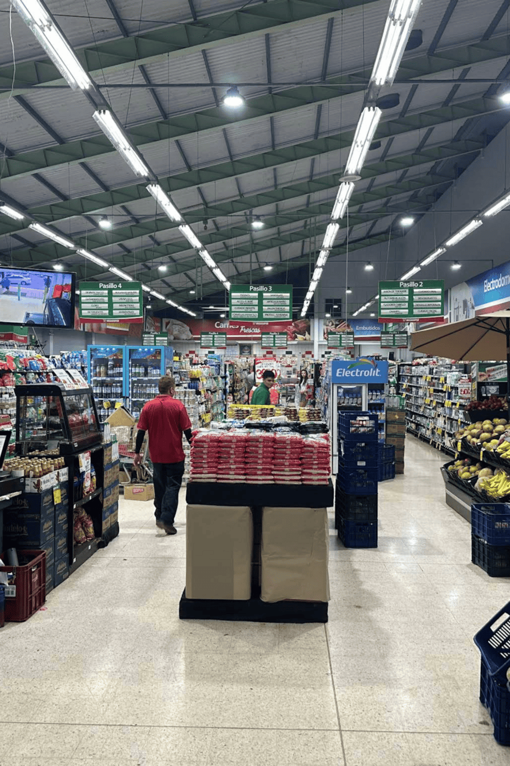 Fresh produce and grocery items inside a well-lit supermarket aisle, featuring signage and shoppers.