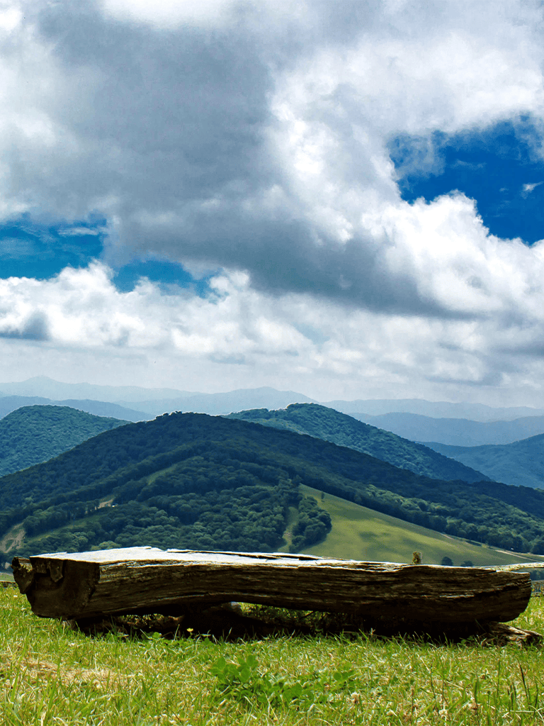 Serene mountain landscape with cloudy sky, lush green hills, and a rustic wooden bench for nature relaxation.