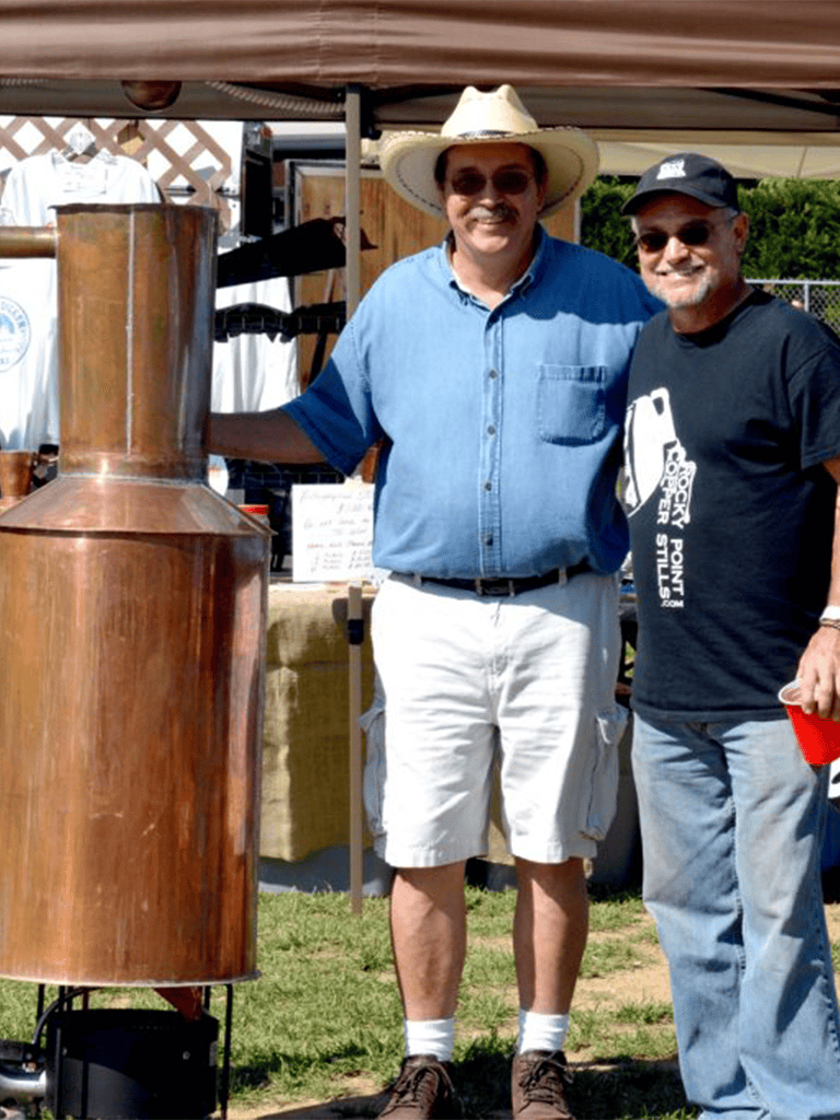 Copper still at outdoor event for distilling spirits, with two smiling men enjoying the occasion.