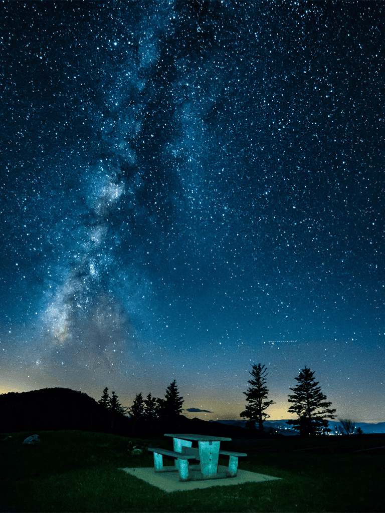 Starry night sky with Milky Way over mountain landscape and outdoor picnic table, lasting astronomical view.