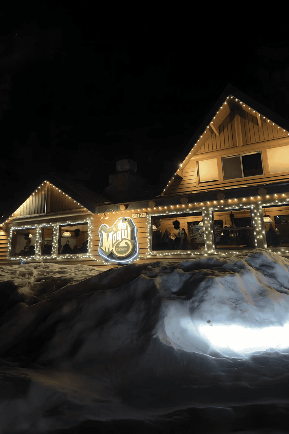 Cozy mountain restaurant illuminated at night with string lights and snow in the foreground.