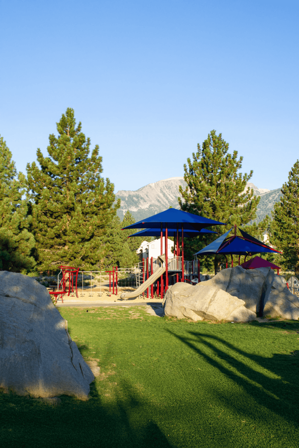 Playground with slides, shaded benches, trees, and mountain background in a scenic park.